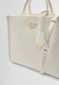 Borsa a tracolla in pelle sintetica bianca con design trapuntato, logo dorato, tracolla staccabile e doppi manici. Presenta dettagli in metallo dorato lucido.