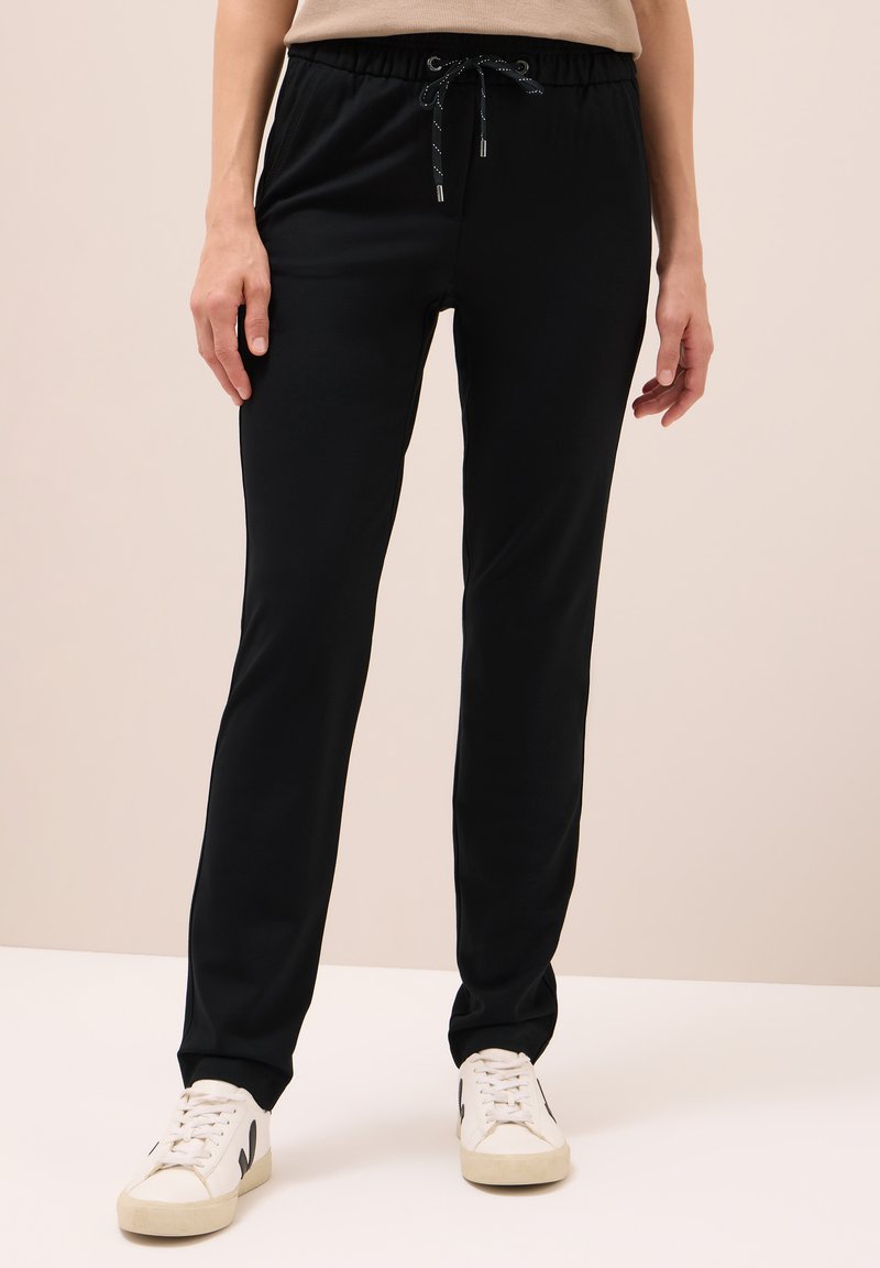 Person, der schwarze Jogginghosen und weiße Sneakers trägt, steht vor einem schlichten hellen Hintergrund.