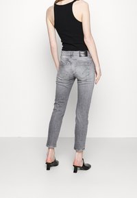 Mujer con jeans ajustados grises, blusa negra sin mangas y mules negros de tacón, de pie de espaldas a la cámara sobre un fondo blanco liso.