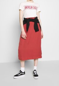 Weißes T-Shirt mit "American Eagle" Grafik, roter Midikleid, schwarzer Pullover um die Taille gebunden, weiße Socken und schwarze Sneakers.