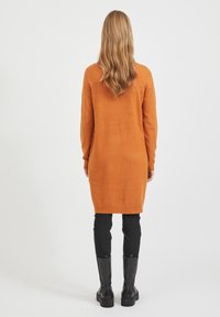Langer, orange gestrickter Pullover mit lockerem Schnitt, ausgestattet mit gerippten Bündchen und Saum, kombiniert mit engen schwarzen Hosen und knielangen schwarzen Stiefeln.