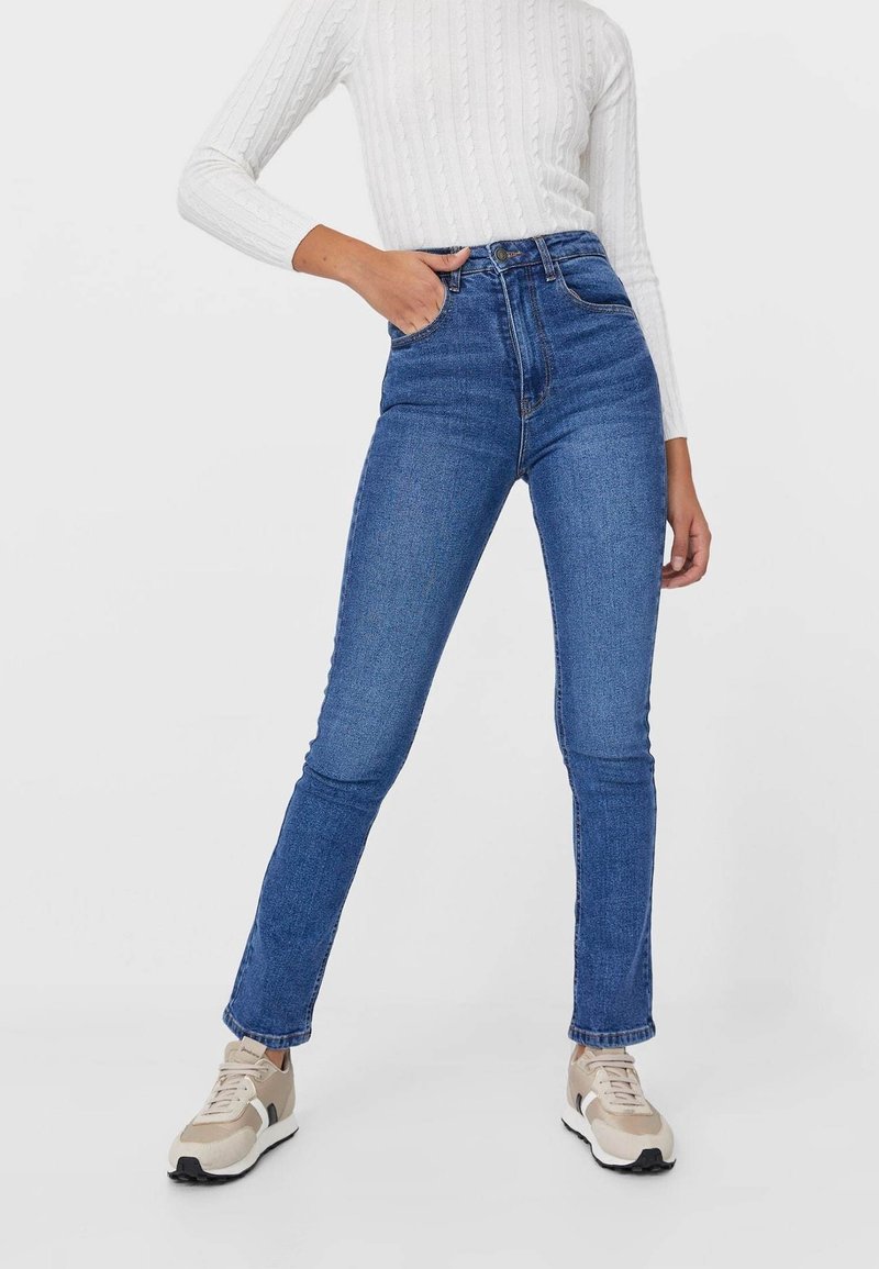 Personne portant un jean bleu taille haute et des baskets beige, une main dans la poche et un pull blanc en maille torsadée.