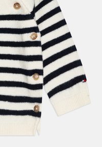 Cardigan rayé crème et marine avec des boutons en bois sur le côté, des poignets côtelés et un petit accent de logo en rouge, blanc et bleu.