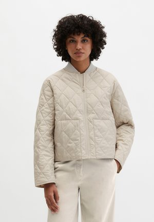 Frau mit lockigem Haar, die eine hellbeige gesteppte Jacke mit Reißverschluss und passende Hose trägt, steht mit einer Hand in der Tasche.