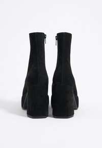 Botines de ante negro con un alto tacón cuadrado, que cuentan con un cierre de cremallera en la parte posterior y un acabado liso y texturizado.