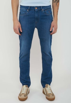 Man die een slim-fit blauwe spijkerbroek en beige sneakers draagt, staand tegen een effen lichte achtergrond.