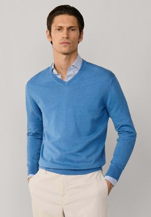 Hombre con suéter azul de cuello en V sobre una camisa de rayas y pantalones beige, con las manos en los bolsillos, de pie contra un fondo claro y liso.