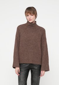 Pull en tricot marron à col roulé avec une coupe ample, manches côtelées, et un ourlet juste en dessous de la taille, associé à un jean foncé et texturé.