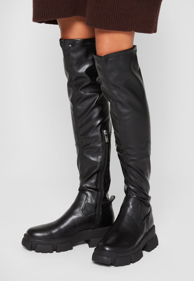 Bottes hautes en cuir noir avec fermeture éclair latérale et semelle épaisse portées avec une jupe marron, photographiées sur un fond uni.