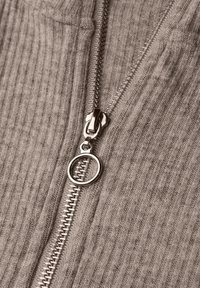 Grå ribbad tyg med en blixtlås i silvermetall. Blixtlåset har ett runt draghandtag som framhäver en texturerad design.