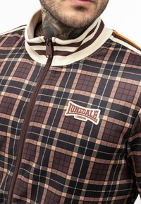 Brown and beige plaid zip-up jacket with cream-colored collar, featuring Lonsdale logo on left chest. Smooth texture, athletic fit.