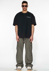 Sort oversized t-shirt med hvidt logo, parret med grønne cargo-bukser med flere lommer og hvide sneakers med brune såler.