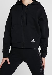 Felpa nera con zip realizzata in materiale morbido, con tasca a marsupio, cappuccio e logo Adidas bianco sul davanti. Design aderente.