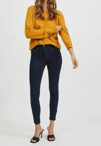 Maglione giallo senape con polsini a coste, abbinato a jeans skinny blu scuro. Scarpe con tacco alto e aperte sulla punta nere completano il look.