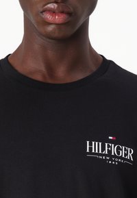 Musta puuvillane t-särk ümarate kaelustega, millel on valge "HILFIGER NEW YORK 1985" logo ja väike punane, valge ja sinine lipu detail.