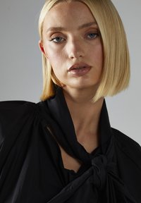 Blusa nera con dettaglio annodato al collo, realizzata in tessuto morbido, con spalle strutturate e scollatura leggermente aperta. Texture liscia.