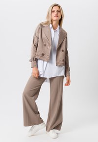 Tan cropped jacket with double-breasted buttons and buckle accents, layered over a white oversized shirt and loose beige pants, paired with white sneakers.