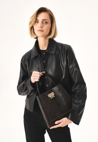 Borsa a mano in pelle nera con un motivo rettile testurizzato di un marrone scuro. Presenta dettagli in oro e manici corti. Il modello indossa una giacca di pelle abbinata.