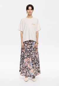Camiseta blanca de gran tamaño con bordado dorado combinada con una falda maxi fluida y estampada en tonos florales pastel. Textura suave, corte relajado.