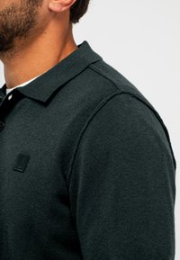 Mann trägt ein dunkelgrünes, langärmeliges Strick-Poloshirt mit geknöpftem Kragen und kleinem quadratischem Logo auf der Brust vor schlichtem Hintergrund.