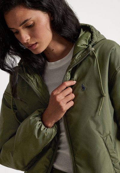 Αδιάβροχο μπουφάν σε χρώμα ελιάς, με κουκούλα, ελαστικές μανσέτες, φερμουάρ στο μπροστινό μέρος και κεντημένο λογότυπο στο στήθος. Με λεία υφή.