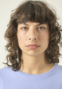 Lockige, dunkle Haare und einen natürlichen Hautton. Trägt ein hellviolettes Shirt. Hat lange, hängende Ohrringe mit klaren Akzenten.