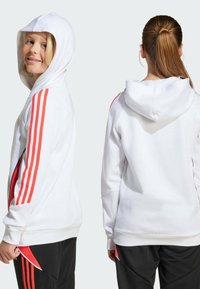 Sweat à capuche blanc avec cordon de serrage, présentant des accents rouges à trois bandes sur les manches et un design sportif. Le tissu semble doux avec une coupe décontractée.