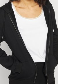 Sort zip-up hættejakke med lommer og snore, lavet af blødt materiale. Bæres over en hvid tanktop med en metallisk kant ved halsen.