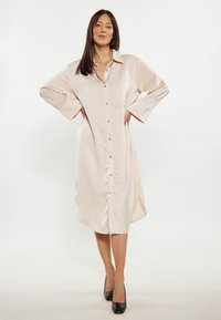 Langes, cremefarbenes Hemdkleid aus glattem Material, mit einer durchgehenden Knopfleiste, Kragen und seitlichen Taschen sowie übergroßen Ärmeln.