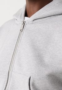 Sudadera gris con cremallera, de textura suave, capucha ajustable y cremallera de metal. Presenta un bolsillo frontal y puños acanalados.
