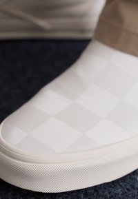 Pantofii albi tip slip-on prezintă un model texturat în carouri cu o finisare netedă, talpă din cauciuc și un design cu vârf rotund.