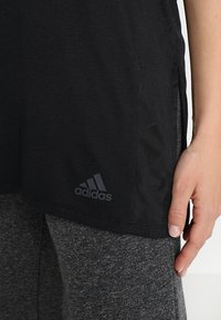 Maglietta sportiva nera in tessuto testurizzato con fessure ai lati, caratterizzata da un logo Adidas grigio in basso a sinistra. Abbinata a leggings grigi.