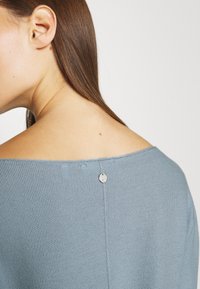 Modrý pletený svetr s designem bez ramínek, s kulatým kovovým tlačítkem na zádech a texturovaným povrchem.