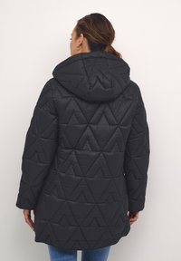Melnas pūkainas jakas ar trīsstūrveida quilted rakstu, kapuci un garām piedurknēm. Izgatavota no spīdīga, gluda materiāla.