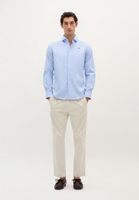 Camicia button-up blu chiaro con un piccolo logo, abbinata a pantaloni crema e scarpe scure, caratterizzata da una vestibilità su misura e una texture liscia.