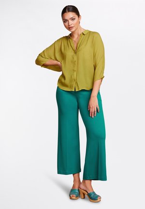 Groene blouse met een knopenlange voorkant en geplooide ontwerp, gecombineerd met losse teal broeken en teal platformklompen. Eenvoudige, vloeiende stoffen.