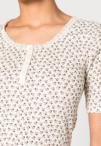 Primer plano de una camiseta blanca de canalé con pequeños estampados florales marrones, cuello con encaje y una tapeta de cuatro botones en la parte superior del torso de una persona.