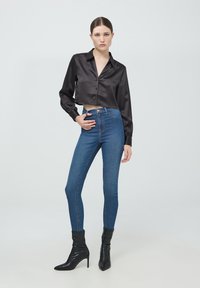 Camicetta nera in satin corta con bottoni, abbinata a jeans skinny blu a vita alta e stivaletti neri con punta.