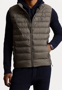 Uppblåsbart väst i olivgrön, med en kviltad design och framdragkedja; bärs över en marinblå stickad hoodie med ribbat mönster.