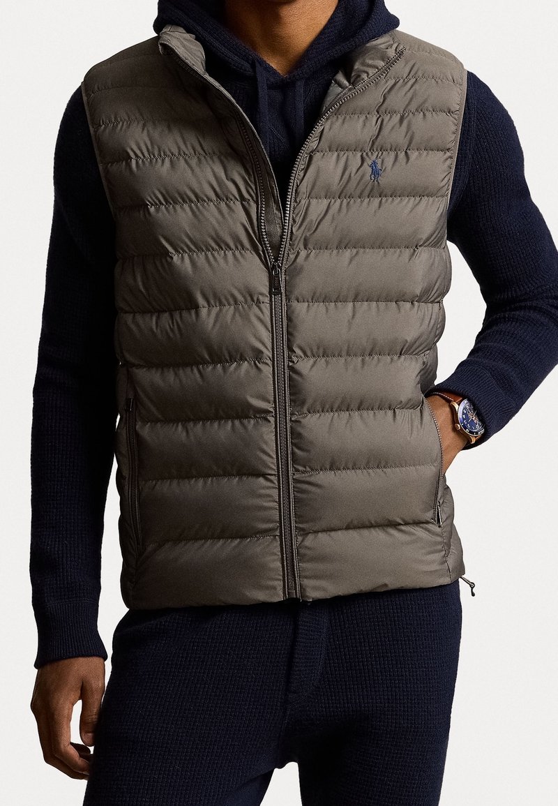 Uppblåsbart väst i olivgrön, med en kviltad design och framdragkedja; bärs över en marinblå stickad hoodie med ribbat mönster.