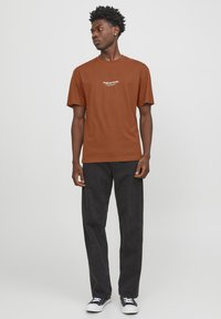 Camiseta de algodón marrón con mangas cortas y logo de texto blanco. Combinada con jeans oscuros y zapatillas negras con acentos blancos.