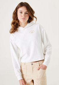 Sudadera blanca recortada con un bolsillo canguro en la parte frontal, que presenta un pequeño gráfico de sol beige en el pecho. Fabricada con un tejido suave y texturizado.