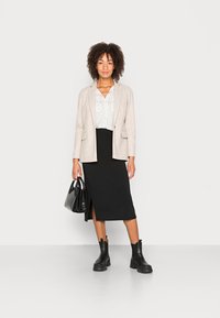 Blazer beige sur une blouse blanche à pois noirs, associé à une jupe midi noire avec une fente sur le côté et des bottines noires.