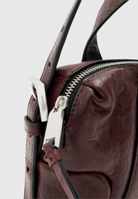 Bordeaux rød lædertaske med sølvfarvet lynlås, tekstureret overflade og en sort justerbar rem. Har metal hardware accenter.