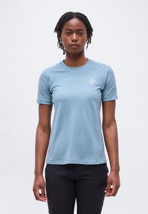 Mlada oseba s kratkimi dreadlocks lasmi, oblečena v svetlo modro srajco s kratkimi rokavi in črne hlače, stoji pred belim ozadjem.