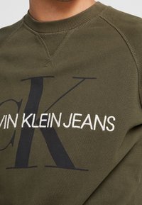Olive green sweatshirt with large black "CK" and white embroidered "CALVIN KLEIN JEANS" logo on chest, ribbed crew neckline.