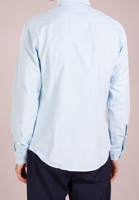 Světle modrá košile s dlouhým rukávem, límcem na knoflíky, texturovanou látkou, jemným leskem a zaobleným spodním lemováním. Má manžety na knoflíky a boční rozparky.