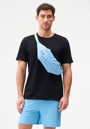 Mann trägt schwarzes T-Shirt, hellblaue Shorts und eine hellblaue Umhängetasche vor einem schlichten weißen Hintergrund.