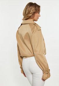 Vaaleanbeige satiinitakki, jossa on korkea kaulus, vyölliset hihat ja muotoiltu istuvuus. Yhdistetty valkoisiin slim fit -housuihin, jotka korostavat tyylikästä muotoilua.