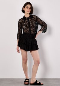 Top de encaje negro con patrones hexagonales, mangas tres cuartos y botones en el frente, combinado con una falda negra con volantes y sandalias planas.